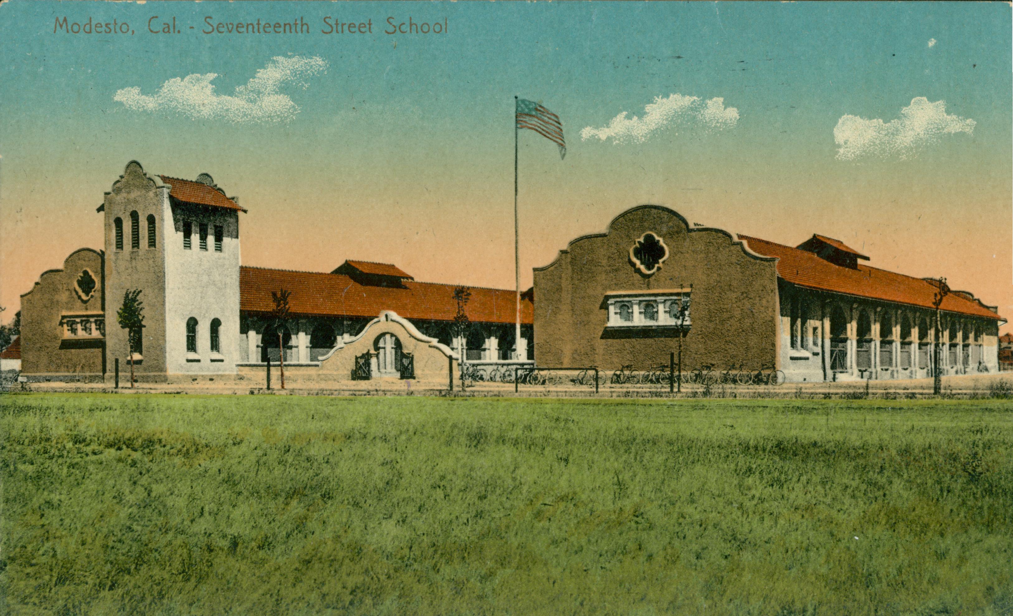 Shows a corner view of the Seventeenth Street School in the middle of an open field with a flag pole out front